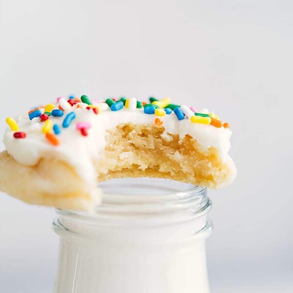 A large bite taken from a delicious soft sugar cookie topped with colorful sprinkles, resting on a container of milk.