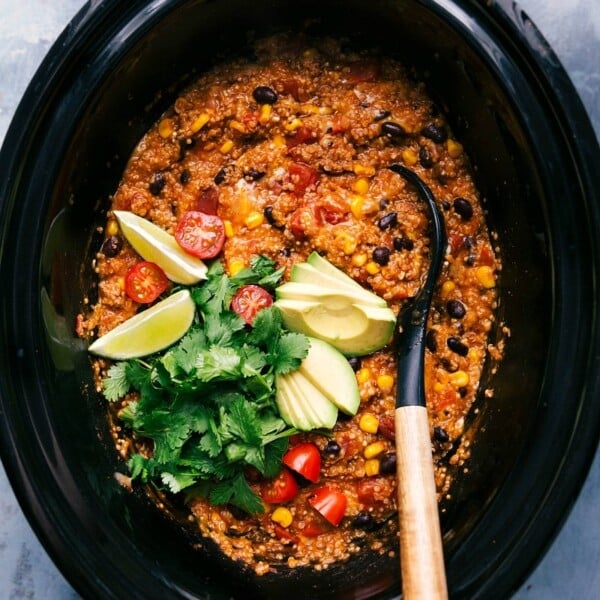 Crockpot quinoa enchilada freshly made, served with a spoon and garnished with cilantro, tomatoes, avocado slices, and lime wedges.