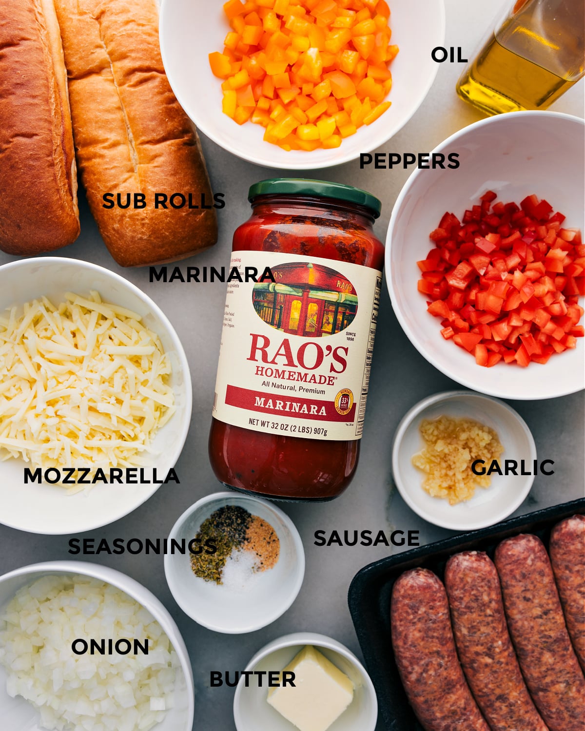 All the ingredients for this recipe prepped out for easy assembly, including peppers, rolls, marinara, mozzarella, onion, seasonings, sausage, garlic, and butter.