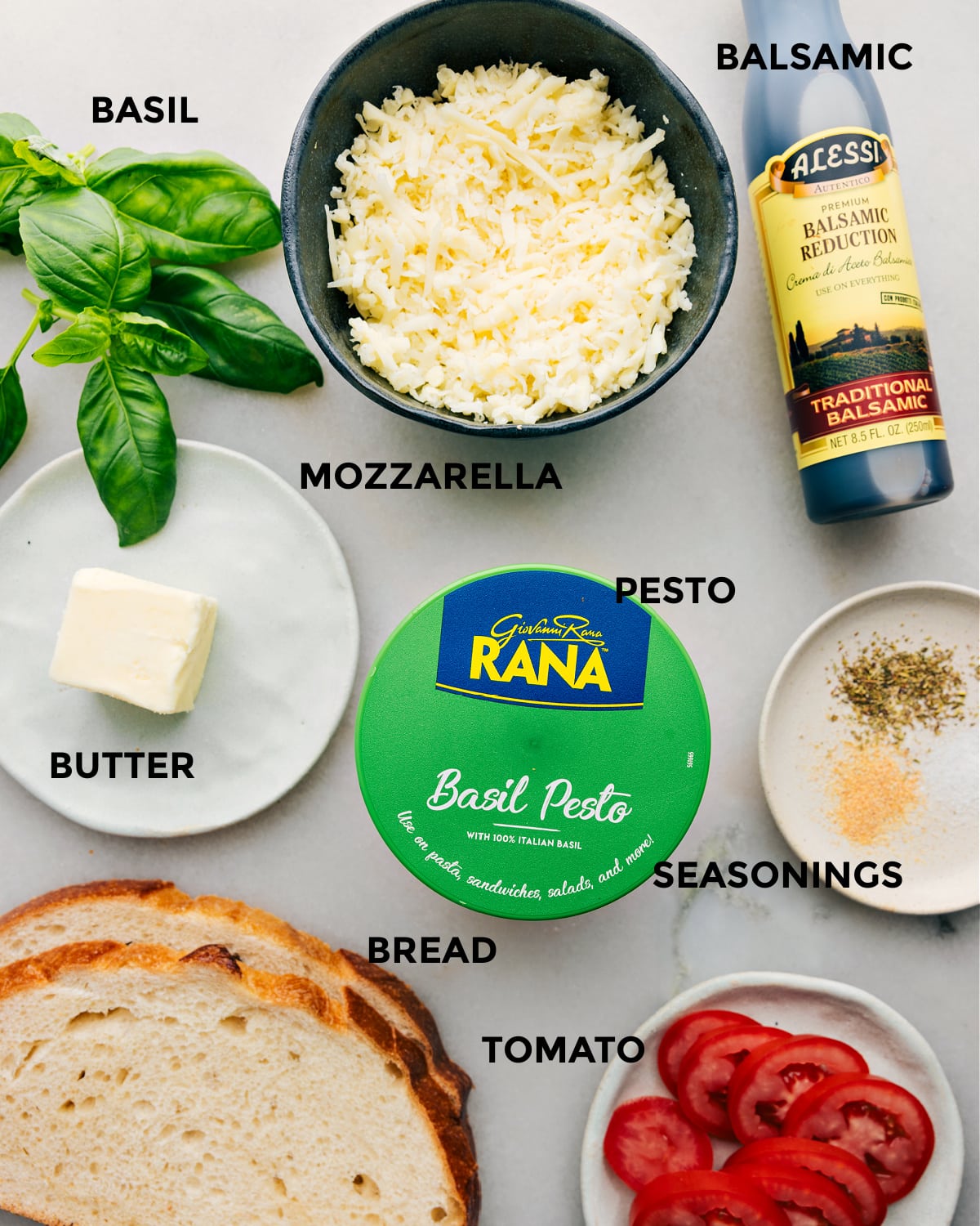 The ingredients in this recipe prepped for easy assembly including basil, balsamic, mozzarella, pesto, butter, seasonings, tomato, and bread.