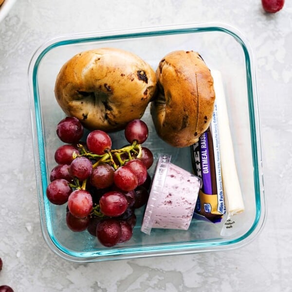 A bagel with a side of cream cheese, fresh grapes, and string cheese in a container, ready to go.