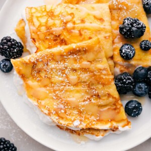 Plate of three crepes with berries and syrup drizzled on top.