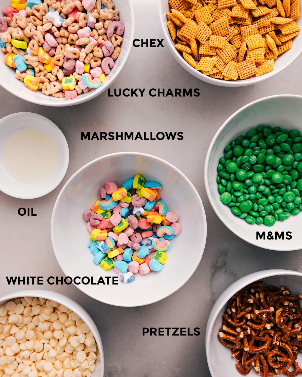 All the ingredients for this recipe are prepped and ready for easy assembly, including Chex, Lucky Charms, M&Ms, marshmallows, oil, chocolate, and pretzels.