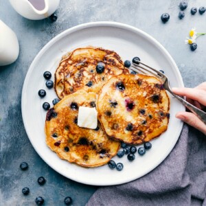 A stack of warm blueberry pancakes on a plate, topped with melting butter and drizzled with syrup, with a fork on the side ready for enjoying.