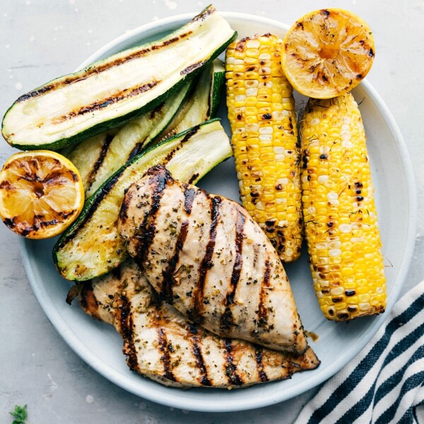 The result of perfectly marinated and grilled meat, served with grilled zucchini, corn, and lemons on the side, creating a mouthwatering meal.