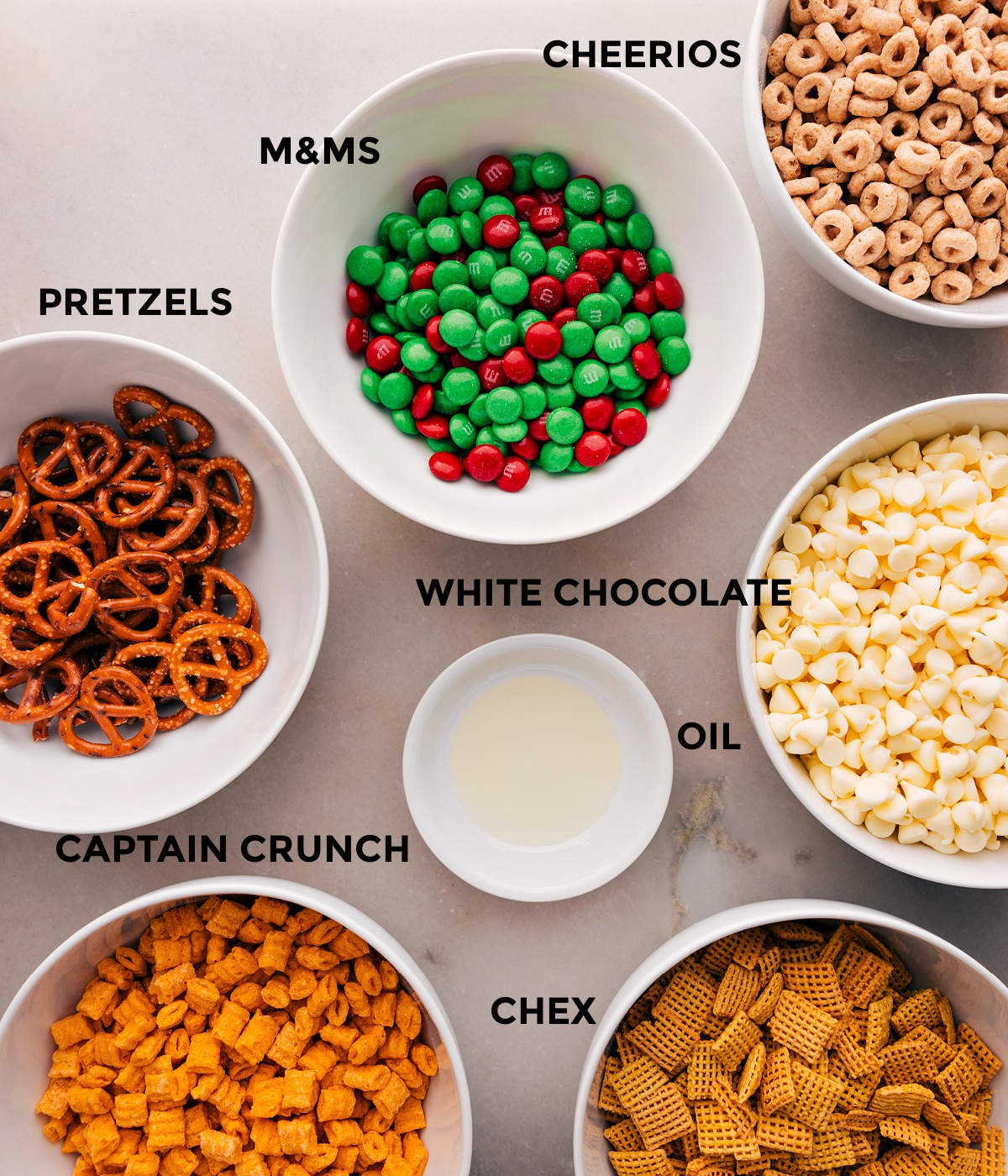 All the ingredients in this recipe are prepped for easy assembly: M&Ms, white chocolate, oil, Chex, Captain Crunch, and pretzels.