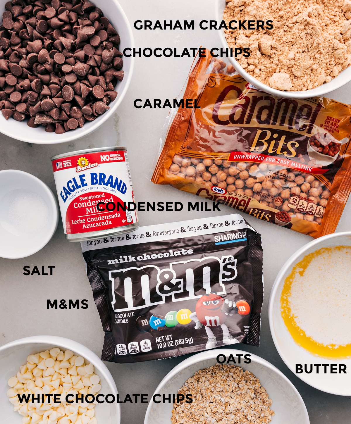 All the ingredients are prepped and ready for easy assembly—graham crackers, caramel bits, condensed milk, chocolate chips, M&Ms, butter, oats, and salt.