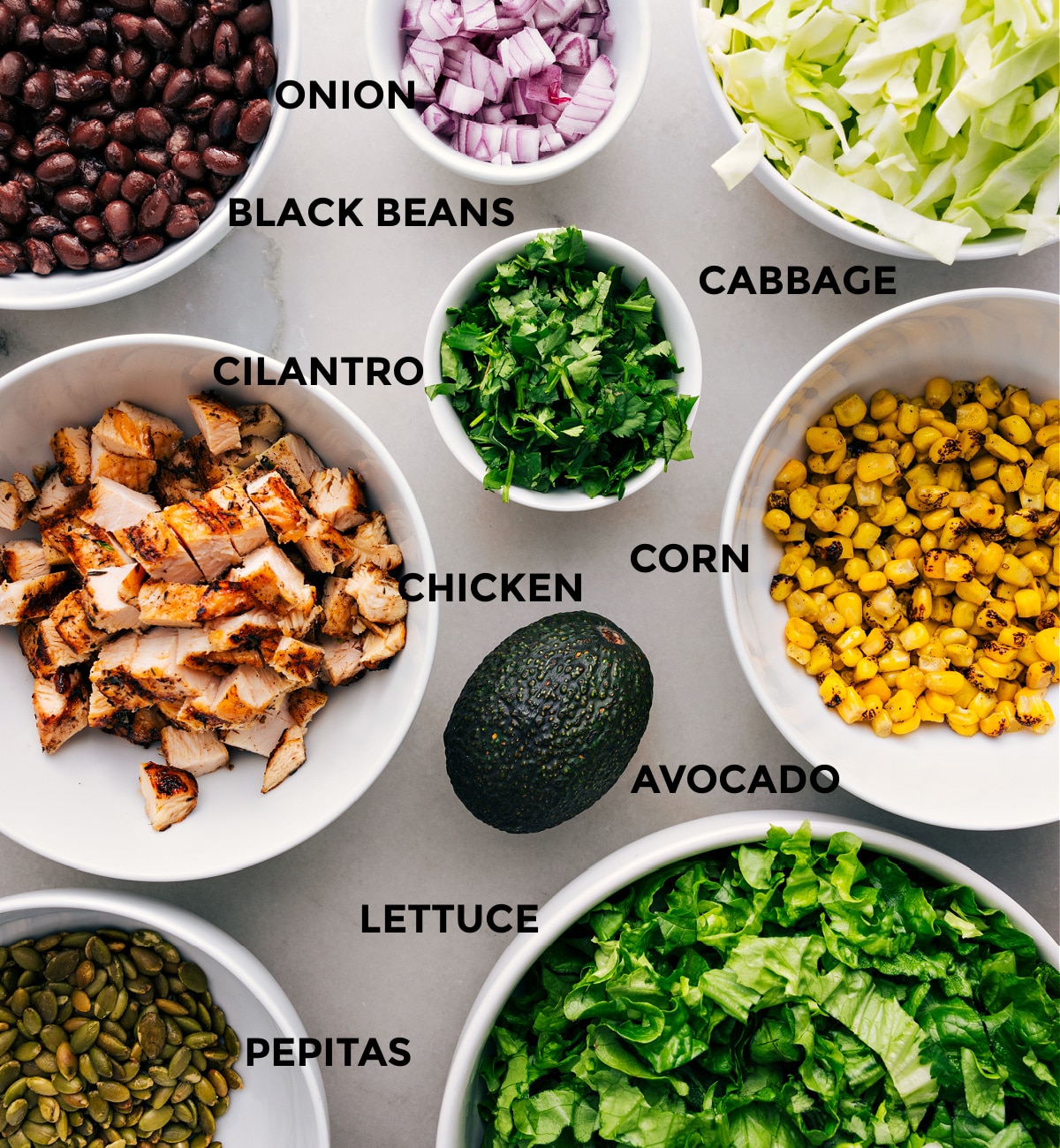 Lettuce and cabbage, pepitas, corn, black beans, avocado, chicken, cilantro, and red onion prepped out for easy assembly of this recipe.