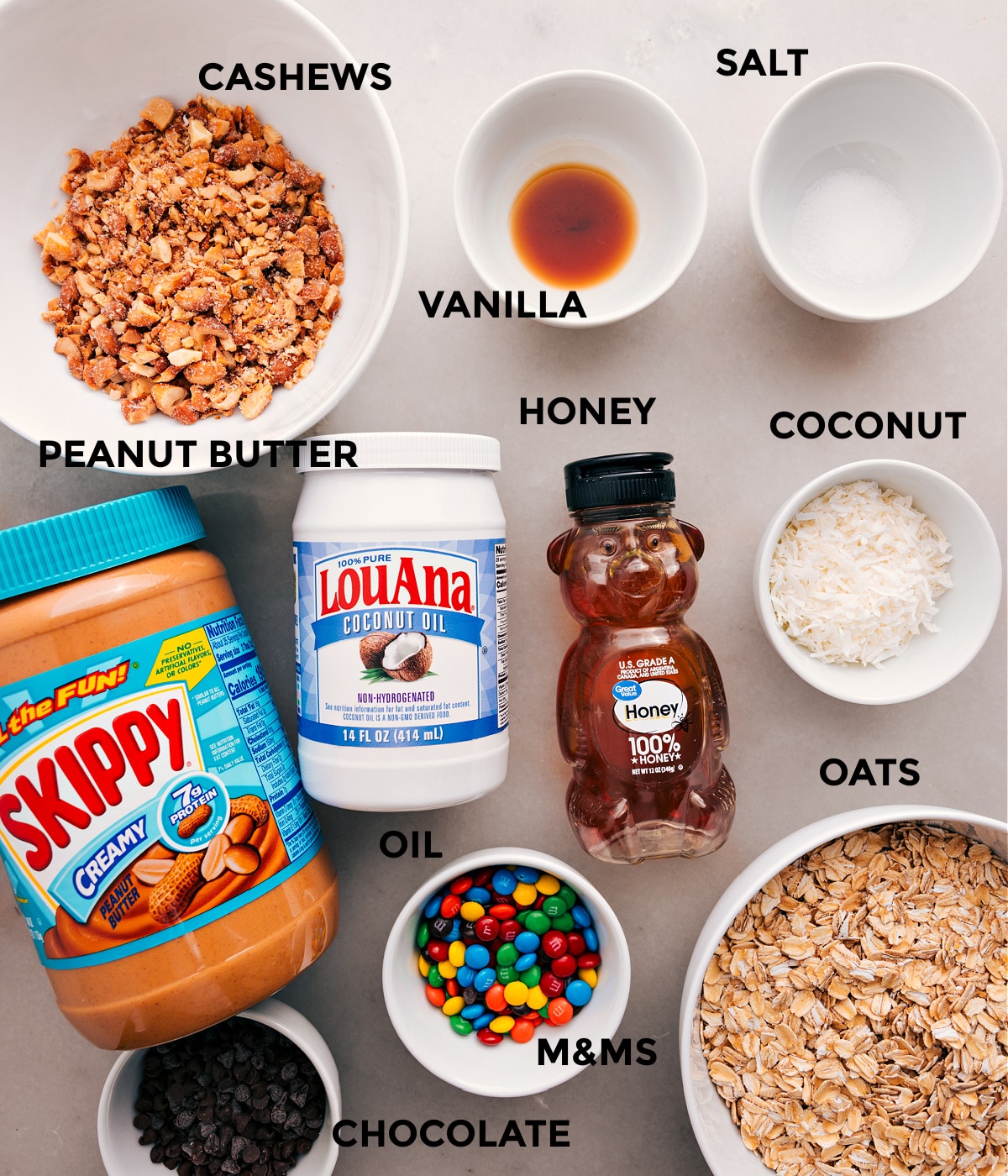 All the ingredients for this recipe are prepped and ready for easy assembly, including cashews, vanilla, salt, coconut, oats, honey, oil, peanut butter, M&Ms, and chocolate.