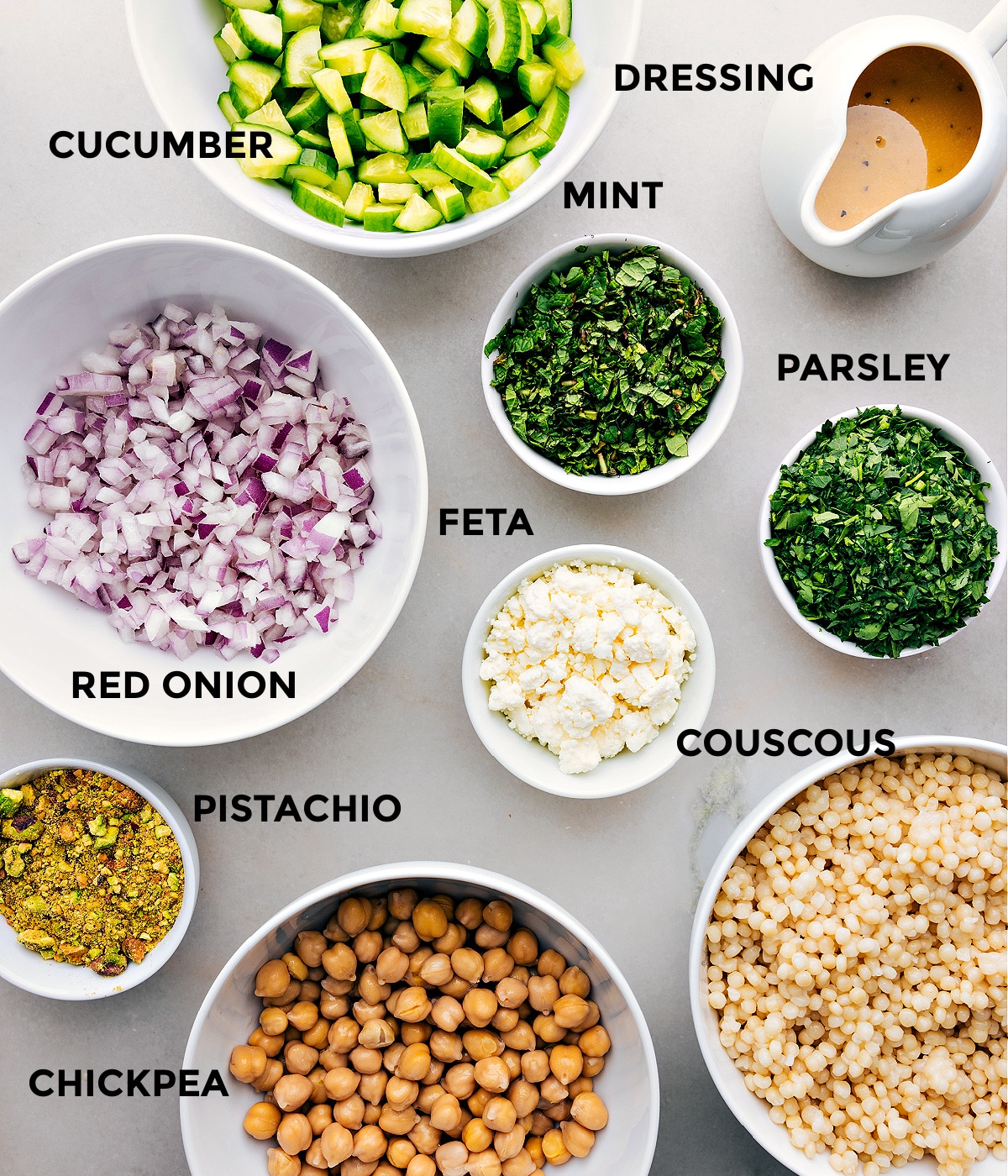 All the ingredients in this recipe are prepped for easy assembly, including cucumbers, mint, dressing, parsley, feta, couscous, pistachios, red onion, and chickpeas.