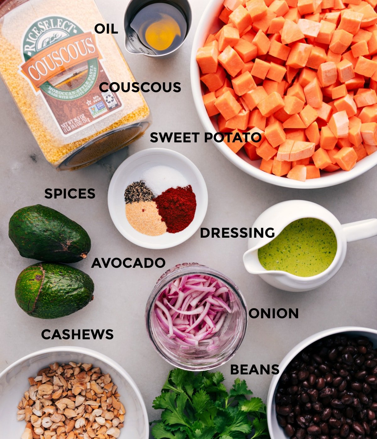 All the ingredients for this recipe are prepped and ready for easy assembly, including sweet potatoes, couscous, dressing, onion, avocado, cashews, and black beans.