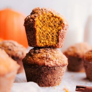 A stack of pumpkin muffins with a warm, golden tone, one at the front displaying a bite taken, revealing the moist interior.