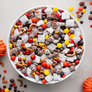 A bowl of Fall Muddy Buddies, showcasing the delightful mix of cereals coated in chocolate and powdered sugar, punctuated by autumn-themed candies and snacks for a festive treat.