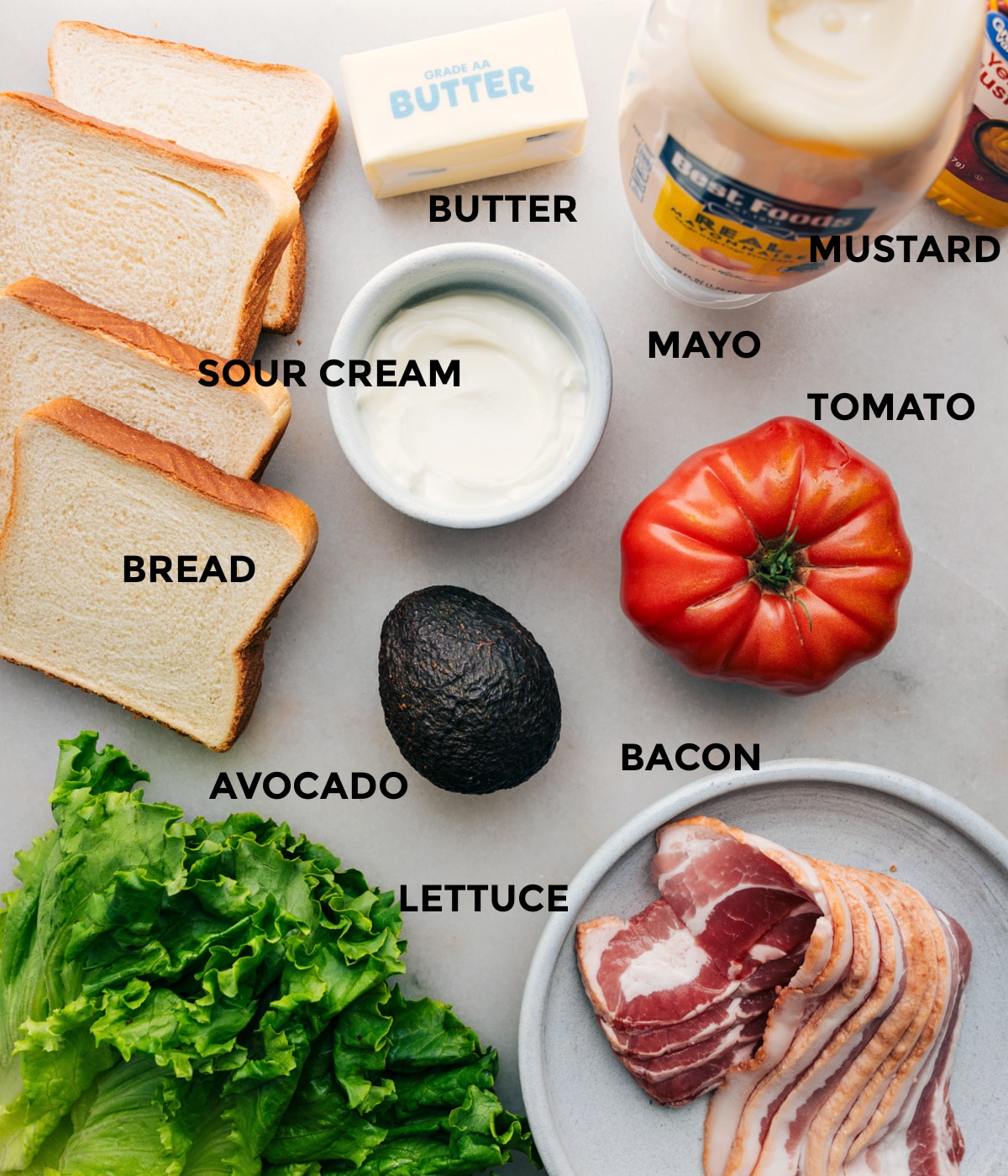 All the ingredients in this recipe including bread, sour cream, butter, mustard, mayo, tomato, bacon, avocado, and lettuce prepped out for easy assembly.