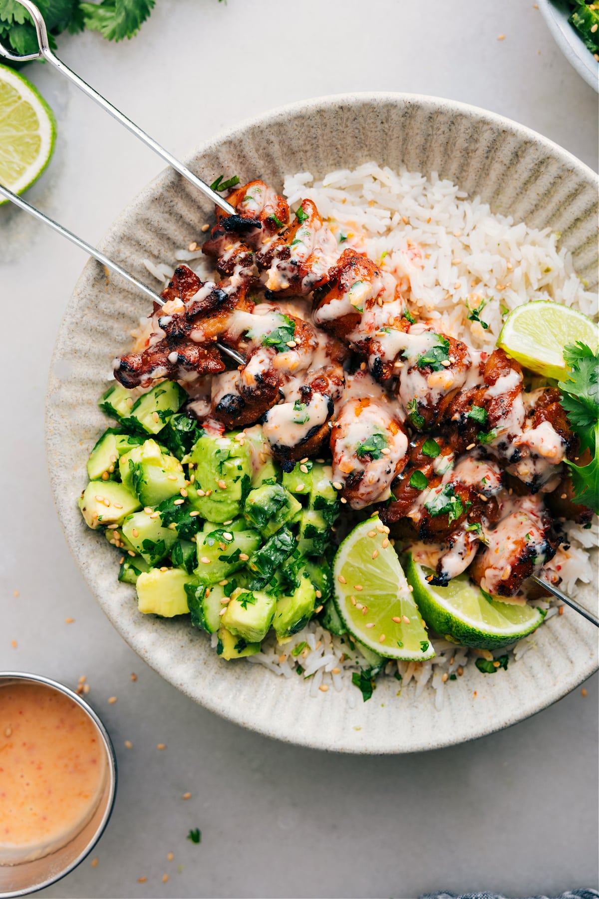 Bang Bang Chicken Skewers sit on a bed of rice, topped with bang bang sauce, and paired with a cucumber avocado salad on the side.