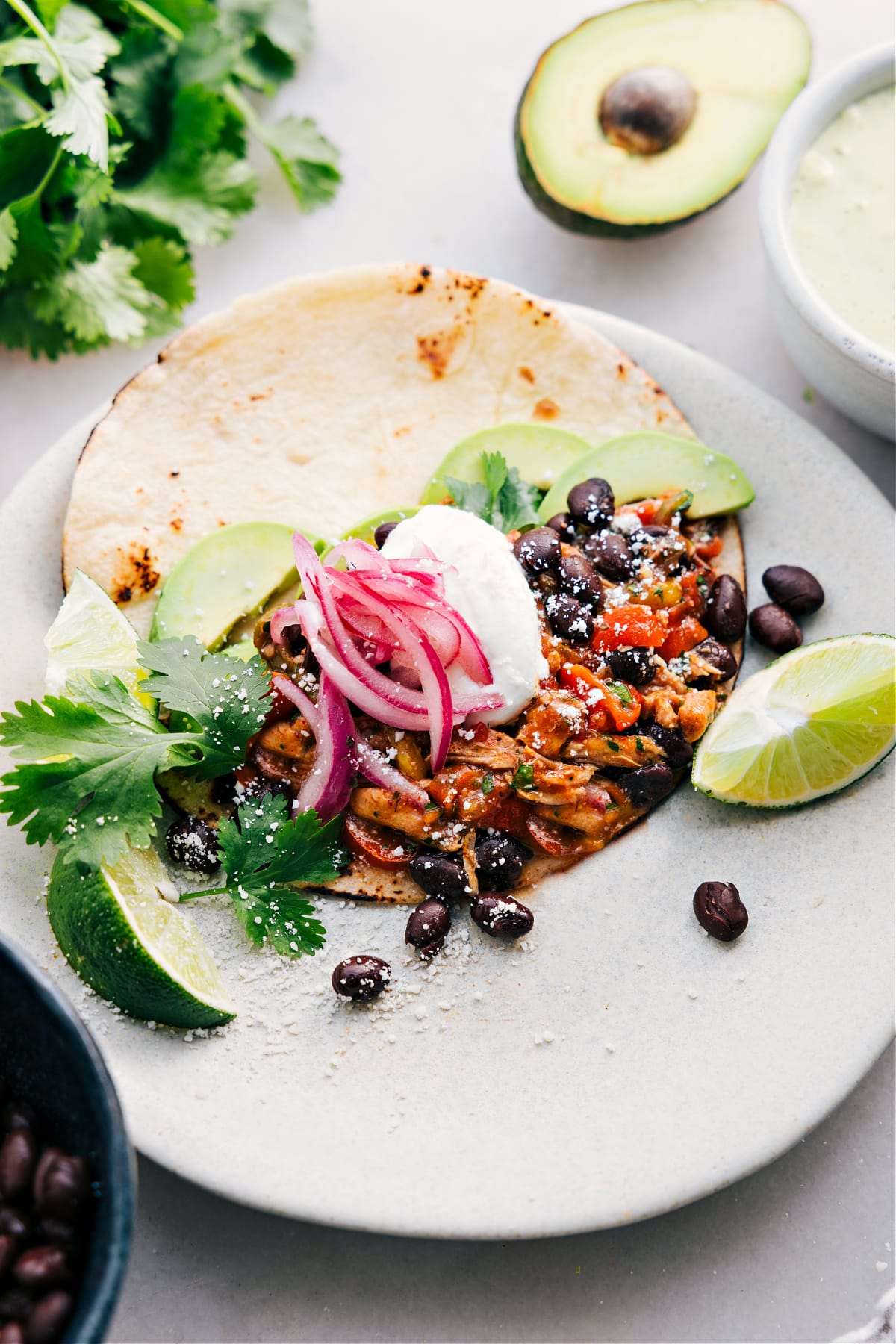 A Crockpot Chicken Fajita assembled in a tortilla with toppings like black beans, onions, sour cream, avocado, cheese, and limes.