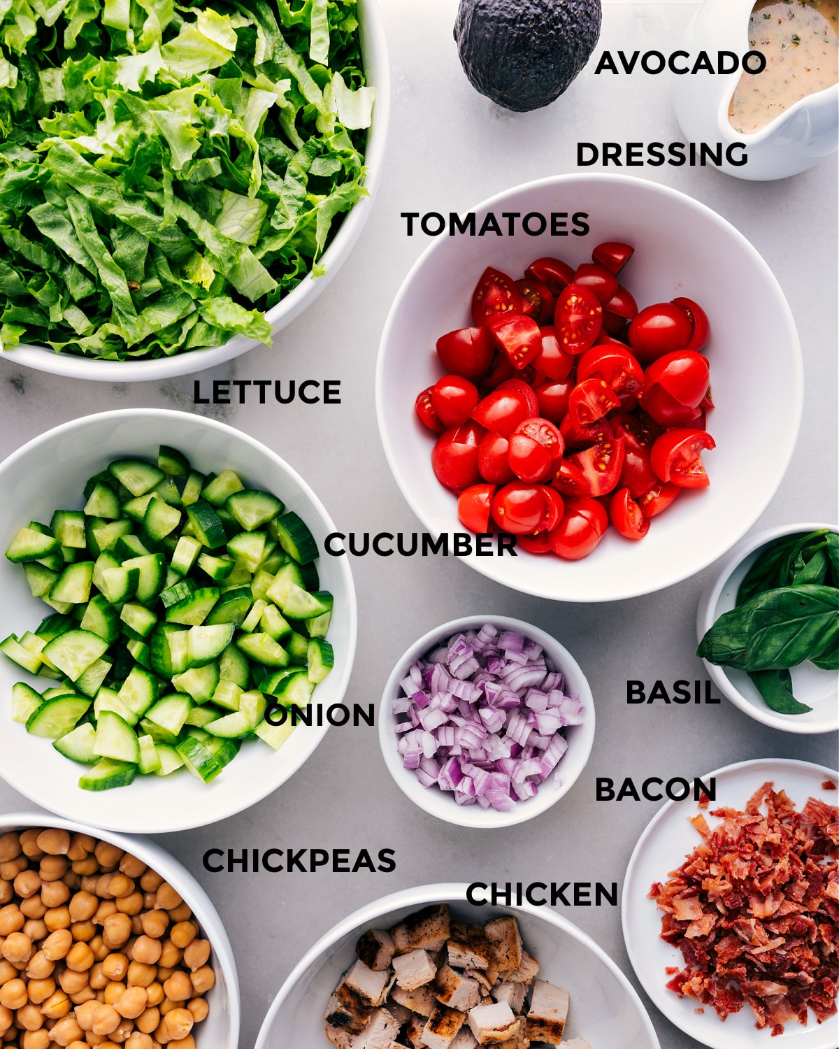 This recipe has everything prepped for easy assembly: tomatoes, avocado, dressing, lettuce, cucumbers, onions, basil, bacon, chicken, and chickpeas.